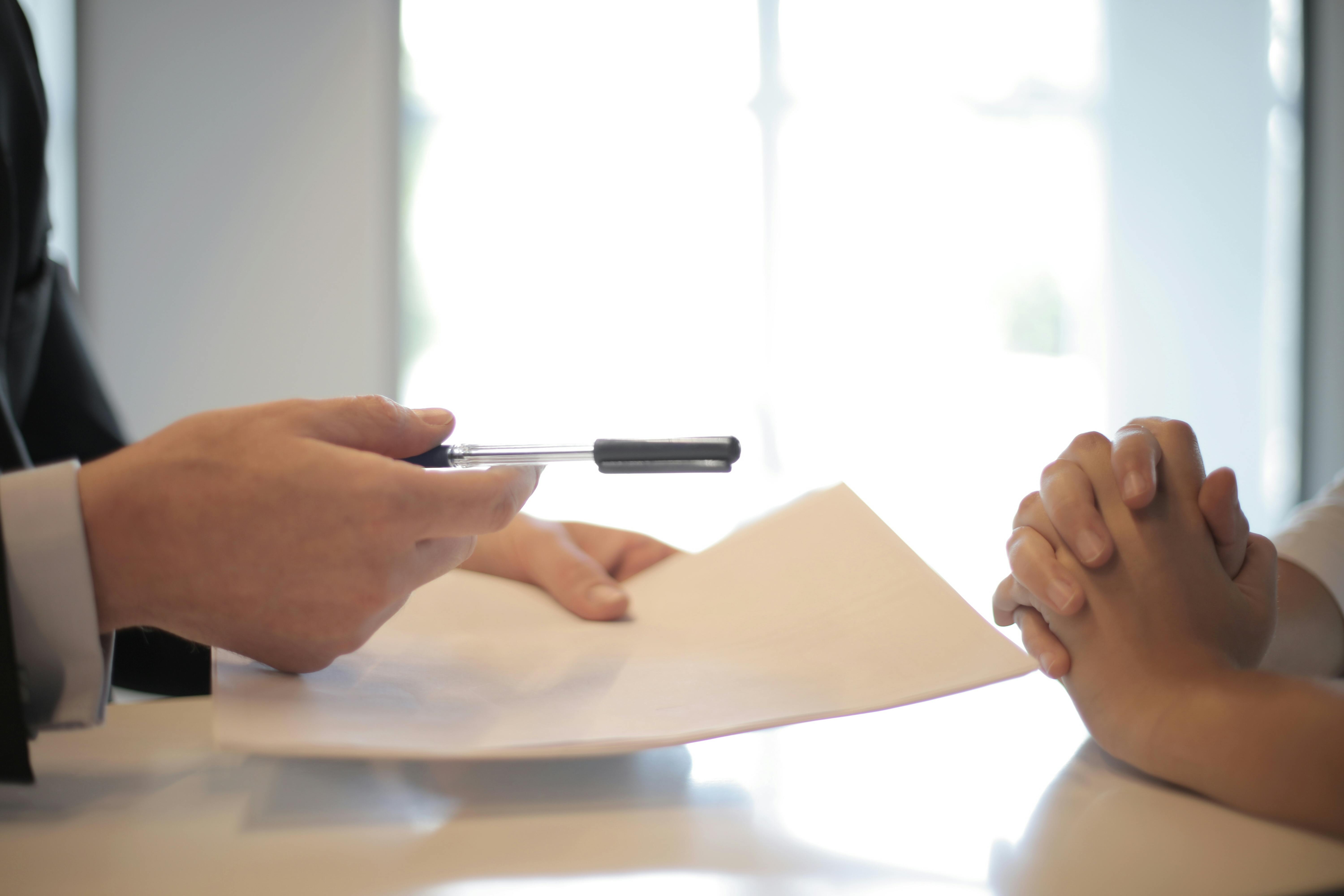 Deux personnes discutent autour d'une table, l'une tenant un stylo et des documents, tandis que l'autre a les mains jointes.