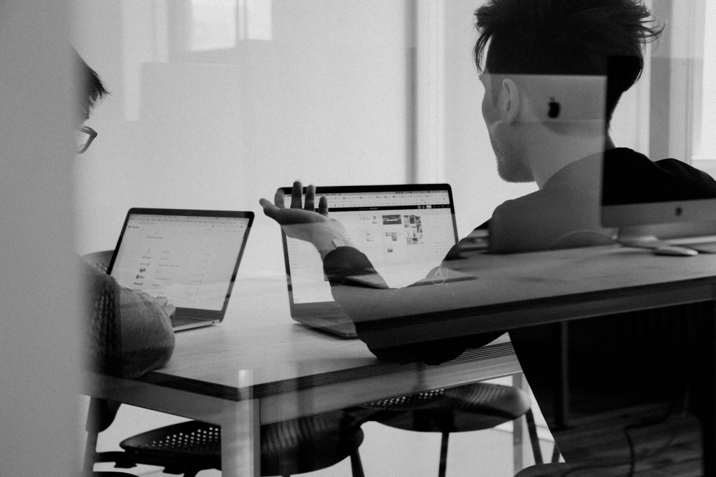 Deux personnes discutant devant des ordinateurs portables dans un environnement de bureau moderne, avec des documents affichés à l'écran.