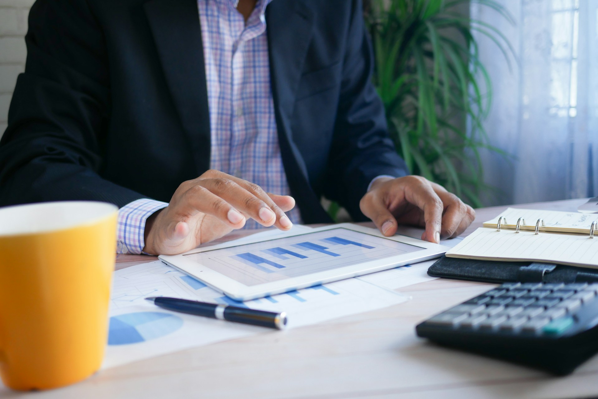 Un professionnel en costume consulte des graphiques sur une tablette, entouré de documents financiers et d'un carnet de notes sur un bureau en bois.
